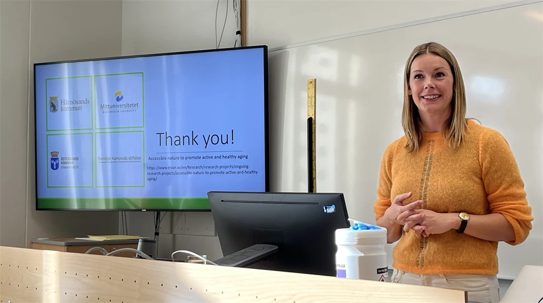 Kvinna med axellångt blondt hår och gul stickat tröja står vid en kateder och ler med en skärm till höger bakom sig