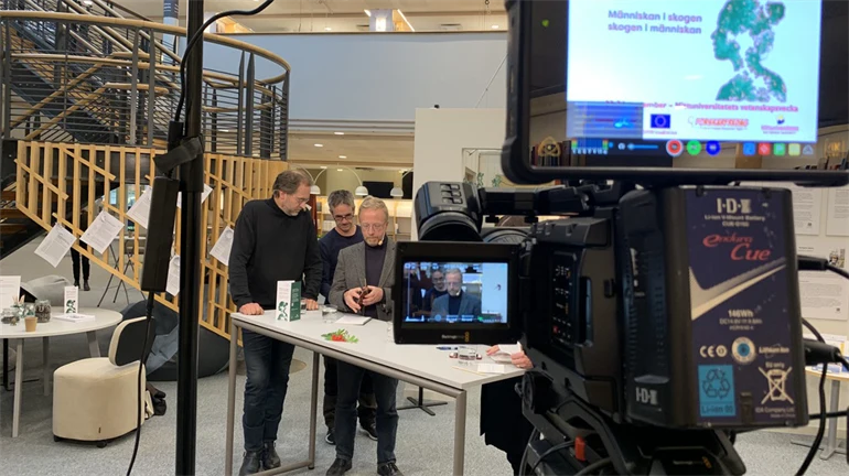 Filmkamera som filmar tre män som samtalar vid ett bord.