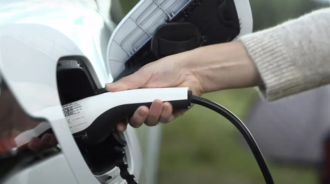 A hand that plugs in a white electric car for charging. Nature and tent can be seen in the background.
