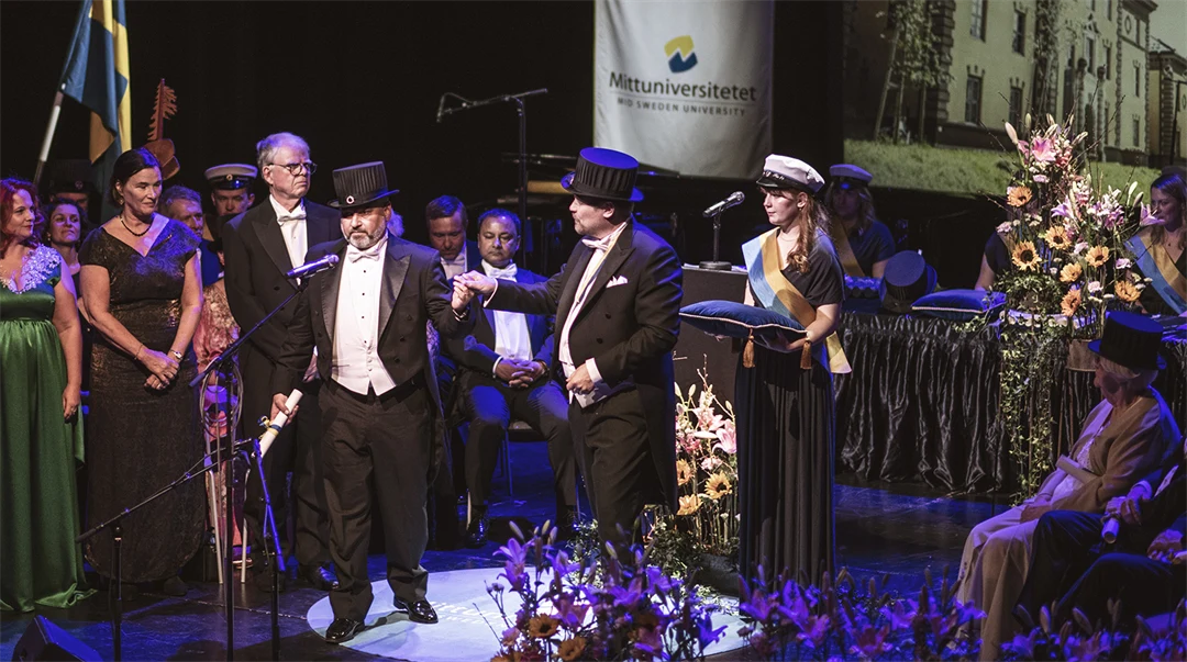 En finklädd person med doktorshatt står i rampljuset på en scen. 