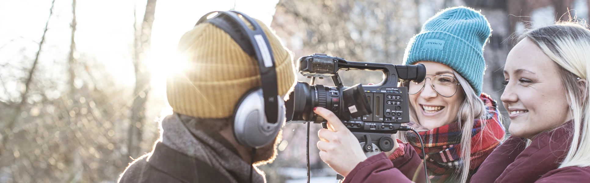 Utomhusbild med en kille med hörlurar som håller i en mikrofon. Bakom en filmkamera står två tjejer.