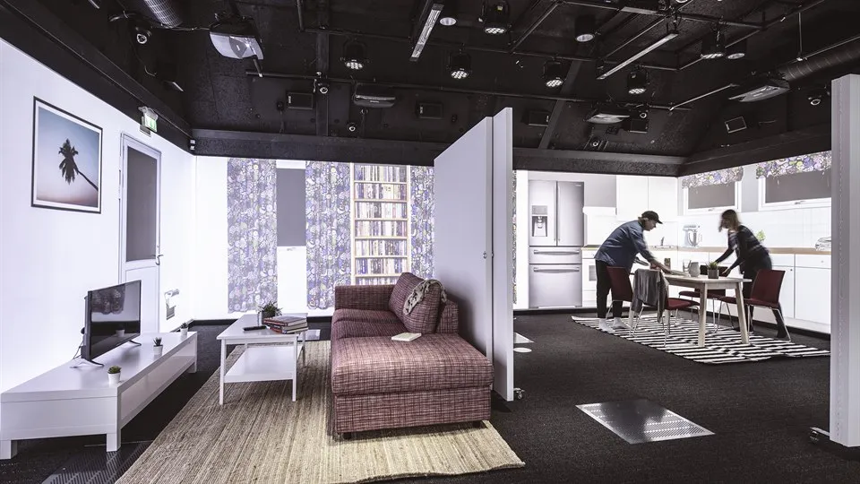 Ett stort rum med svart golv och tak. Rummet ser ut som en lägenhet med mellanväggar, möbler, och projiceringar på väggarna. Två personer står vid köksbordet. I taket finns massor av teknisk utrustning.