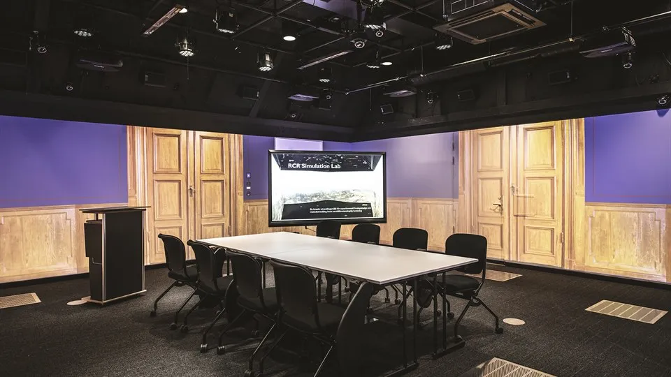 Ett stort rum med svart golv och tak. På väggarna projiceras bilder från ett äldre, pampigt mötesrum. Rummet är möblerat med ett stort bord och åtta stolar, en datorskärm och en talarpulpet. I taket finns massor av teknisk utrustning.