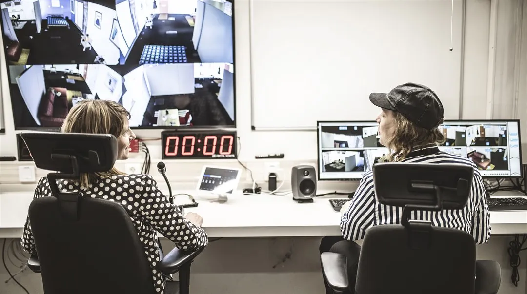 Two people are sitting at a long work table. In front of them are several computer screens, a microphone and other technical equipment.