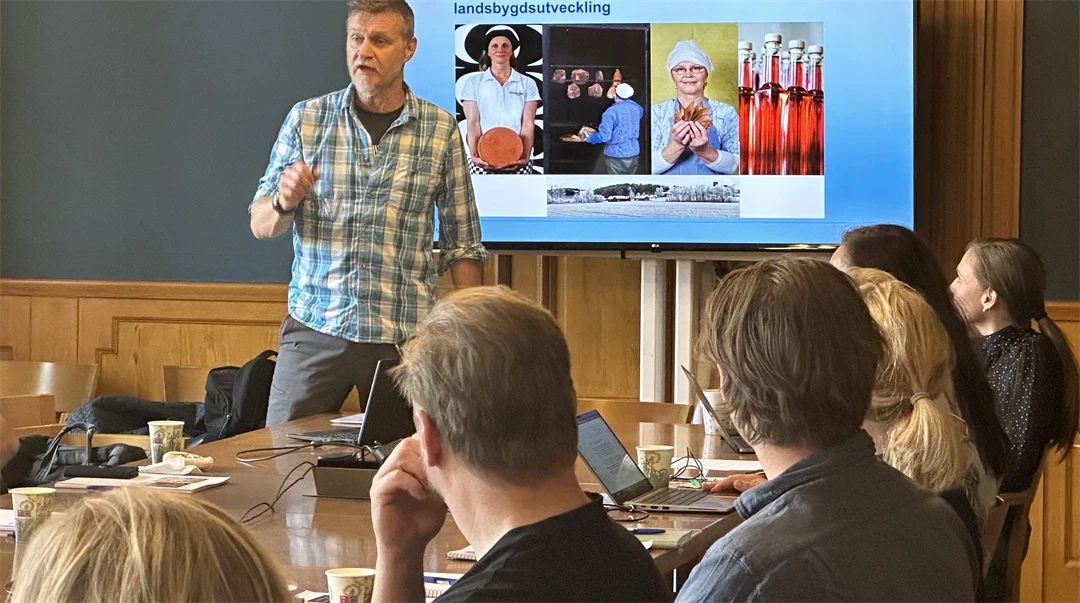 A man in a checked shirt stands in front of the seated audience and presents with a screen behind him.