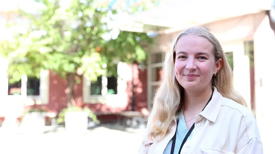 Porträttbild av kvinna med ljust långt hår och vit skjorta. Rött hus och ett grönt träd i bakgrunden. 