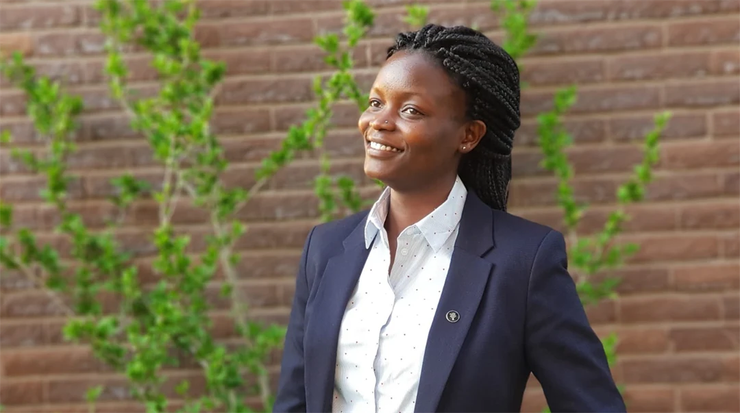 A woman with black hair up, blue jacket and white shirt standing by a brick wall.
