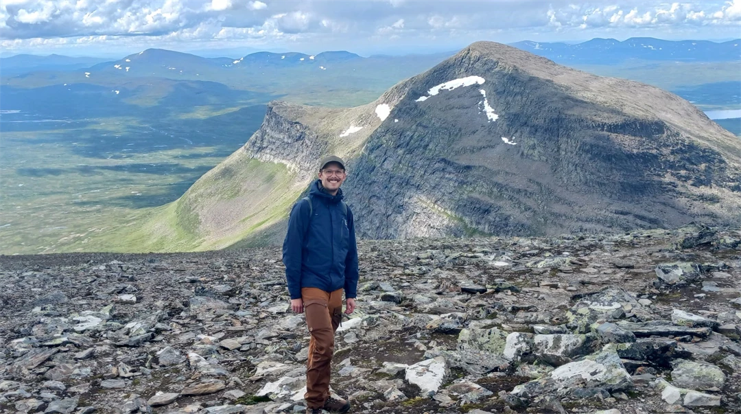 Man som i blå jacka, bruna byxor och keps står utomhus i stenigt landskap med berg i bakgrunden