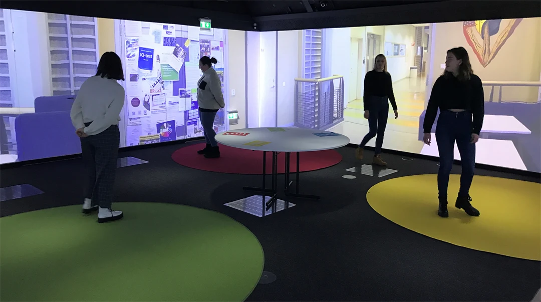 Four people are standing in a room. There are coloured circles on the floor. Some of the people are standing on the coloured circles. Pictures of a school environment are projected on the walls.