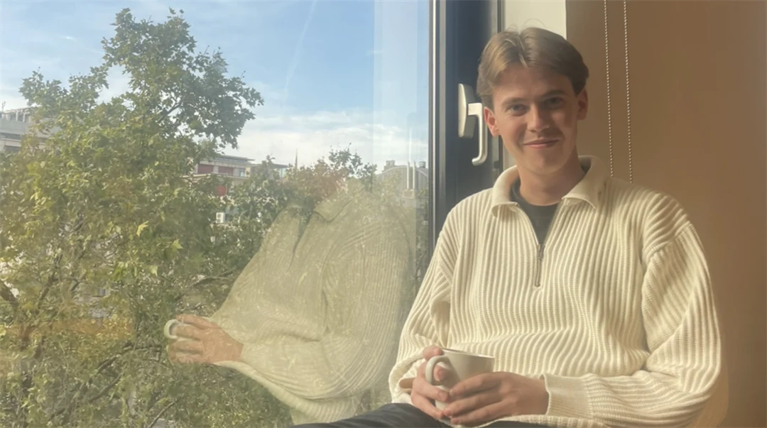 Ung man som sitter i ett fönster med en vit stickad tröja på sig och med en kaffekopp i handen. 