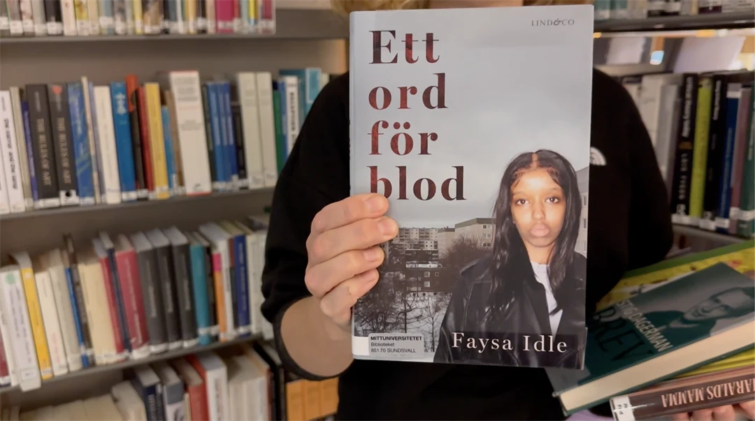 en hand håller upp en bok, bokhyllor i bakgrunden